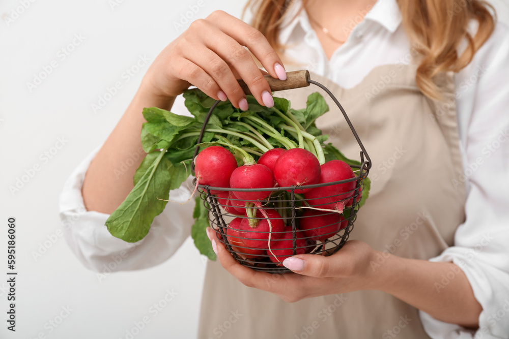 Woman holding basket of ripe radish with green leaves on light background, closeup