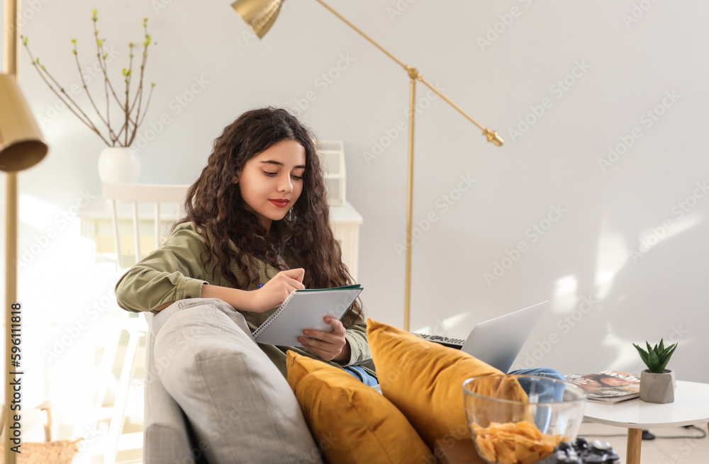 Teenage girl with laptop writing in notebook on sofa at home