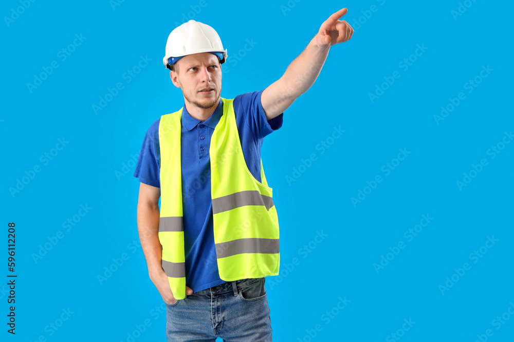 Male worker in vest and hardhat pointing at something on blue background