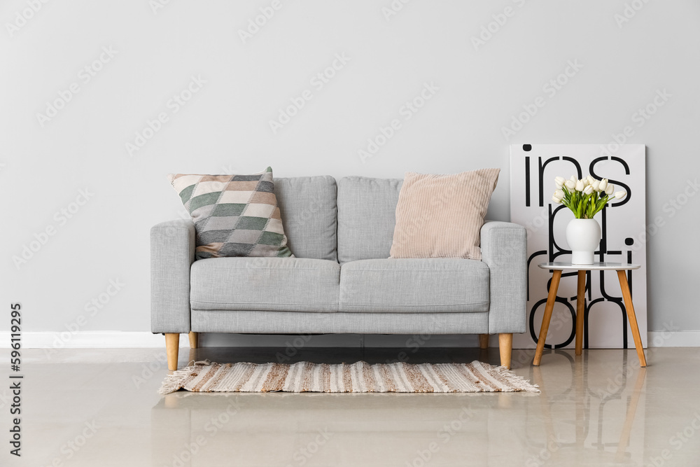 Cozy sofa with cushions and tulip flowers in vase on table near grey wall