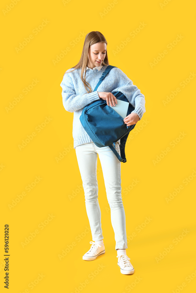 Female student putting copybook into backpack on yellow background