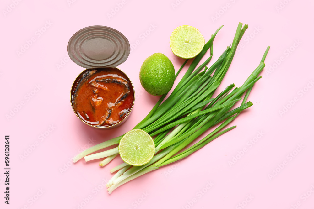 Opened tin can with fish in tomato sauce, lime and scallions on pink background