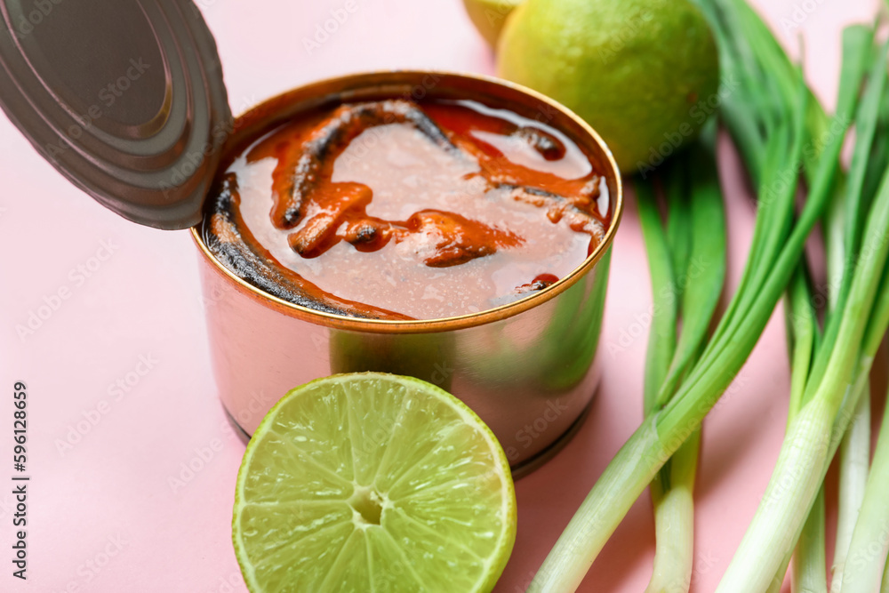 Opened tin can with fish in tomato sauce, lime and scallions on pink background