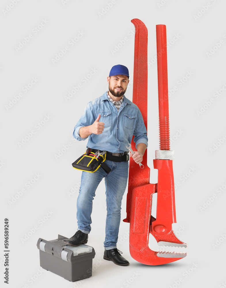 Handsome plumber with big pipe wrench showing thumb-up on light background