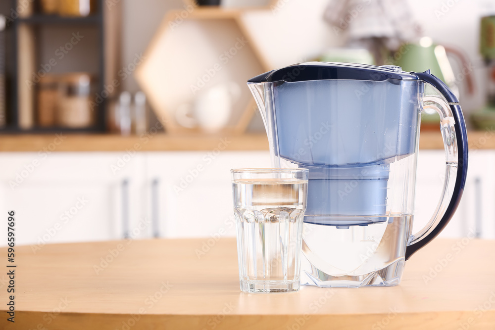 Modern filter jug and glass of water on table in kitchen