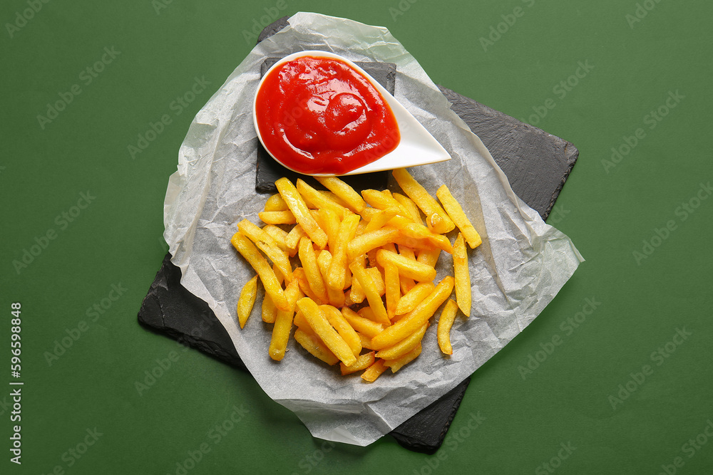 Slate board of tasty french fries and ketchup on green background