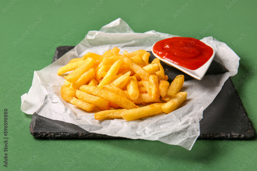 Slate board of tasty french fries and ketchup on green background