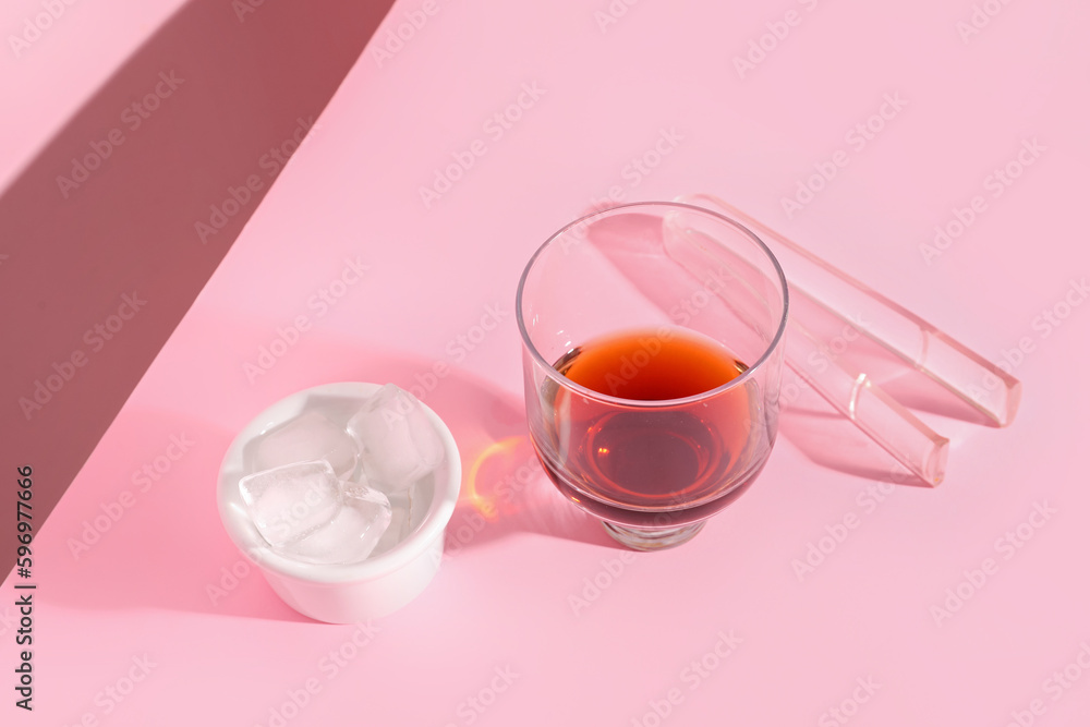 Glass of rum, bowl with ice cubes and tongs on pink background