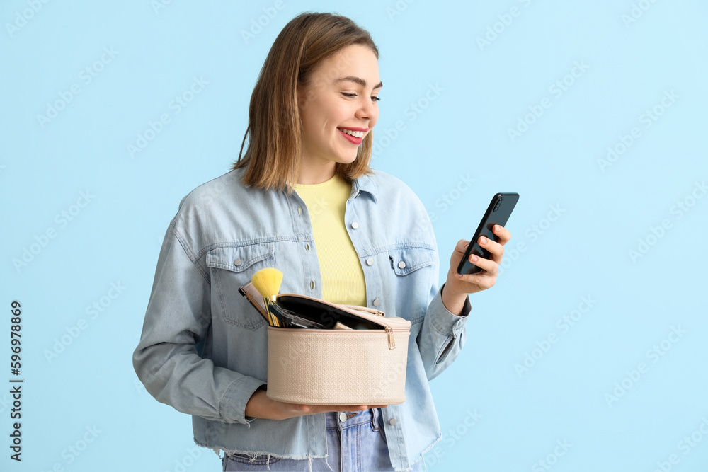 Young woman with cosmetic bag using mobile phone on blue background