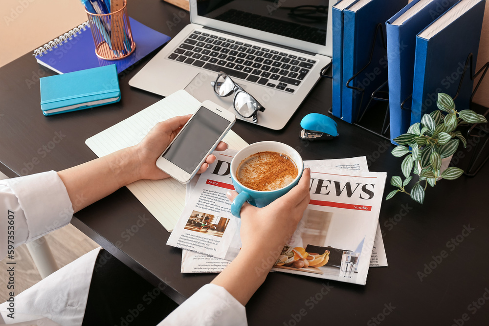 Woman with cup of coffee and mobile phone at table in office, closeup