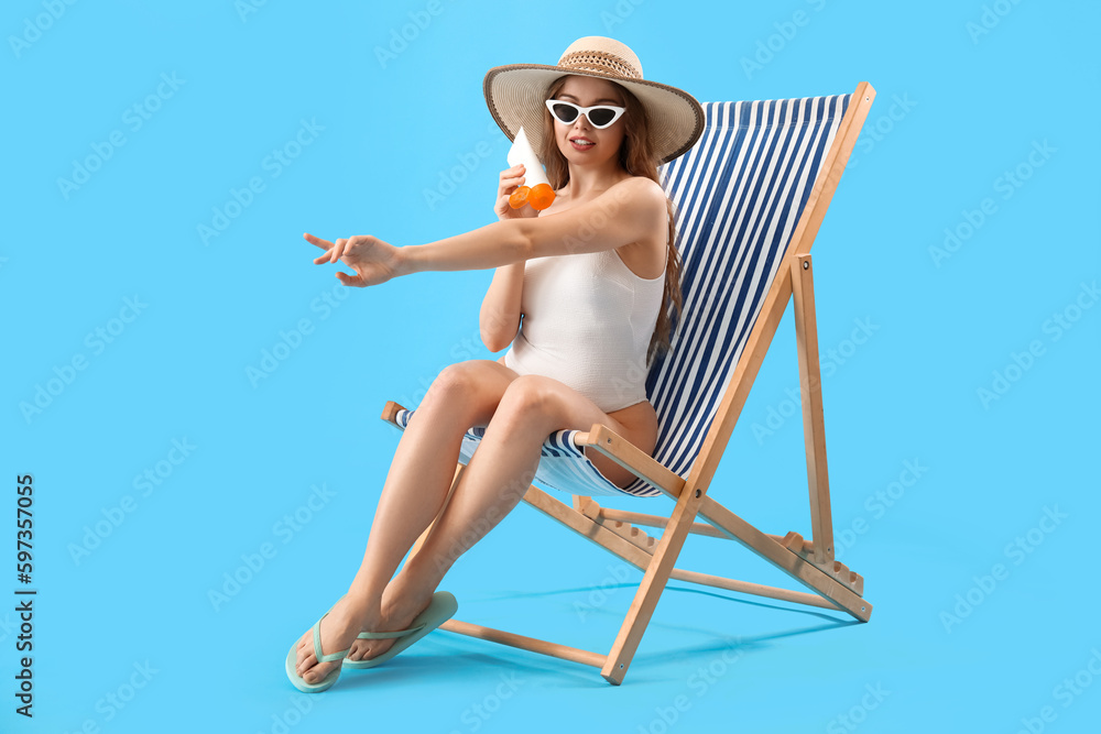 Beautiful young woman applying sunscreen cream while sitting on deck chair against blue background