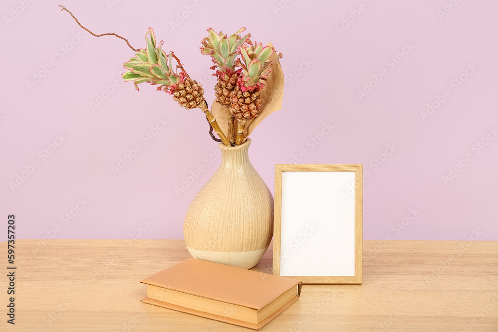 Vase with baby pineapples, book and blank frame on table near lilac wall