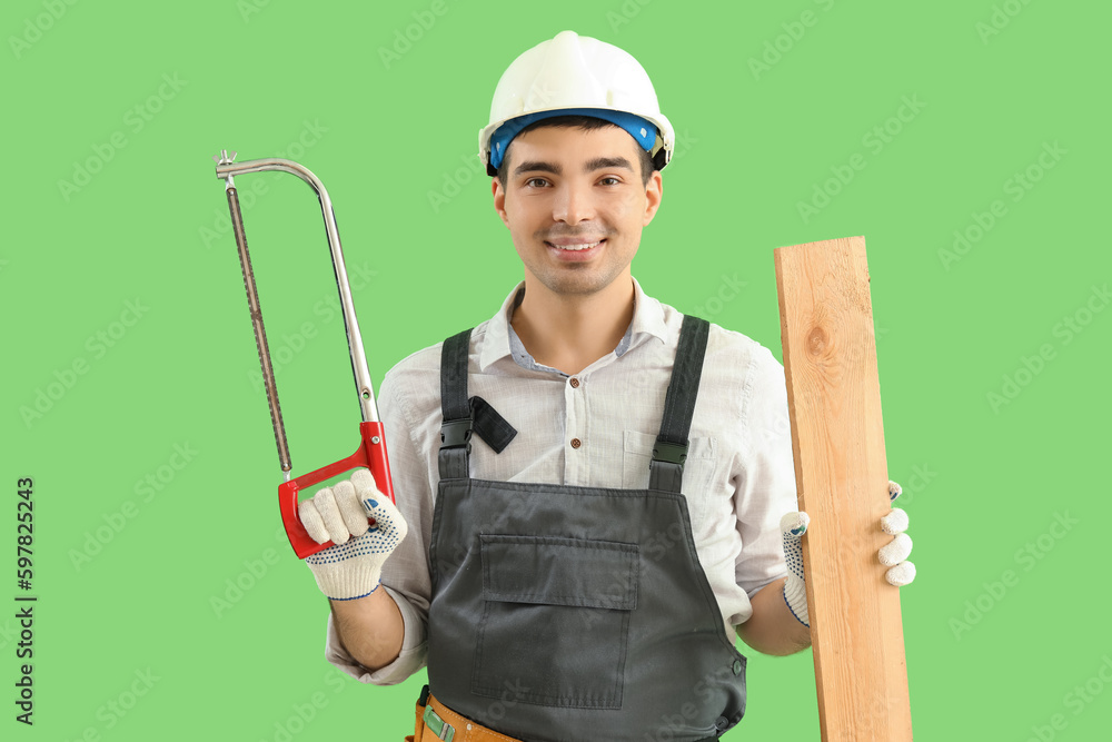 Young carpenter with hacksaw and wooden plank on green background