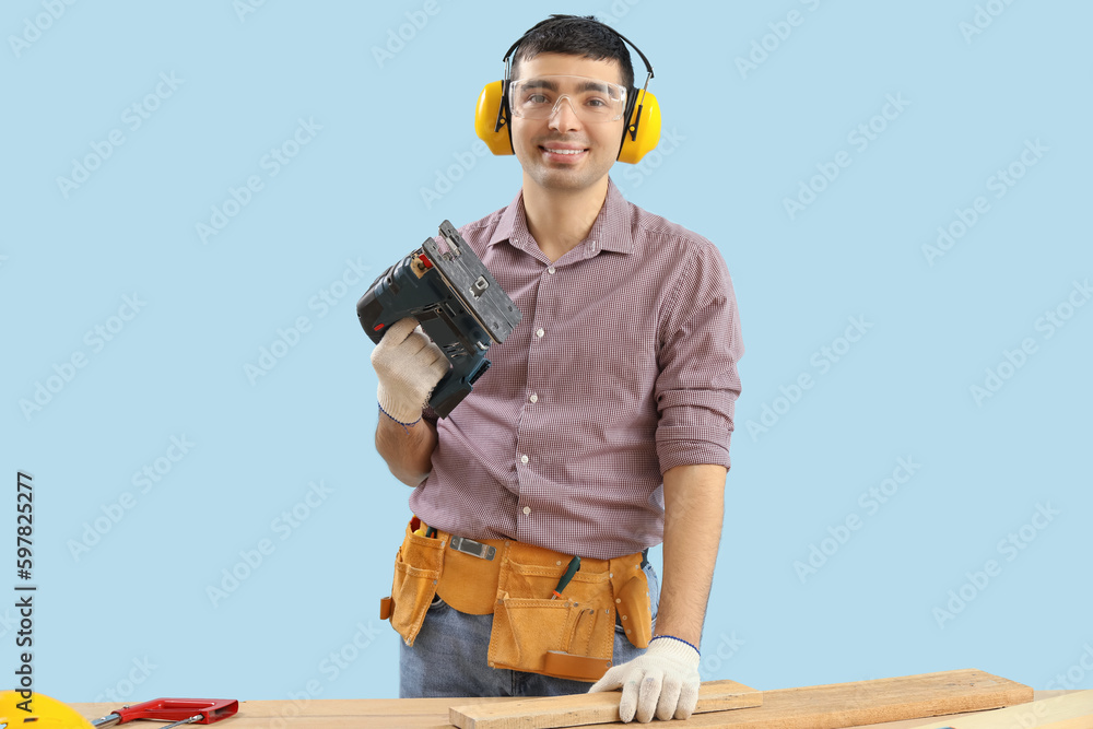 Young carpenter with jigsaw and wooden plank at table on blue background