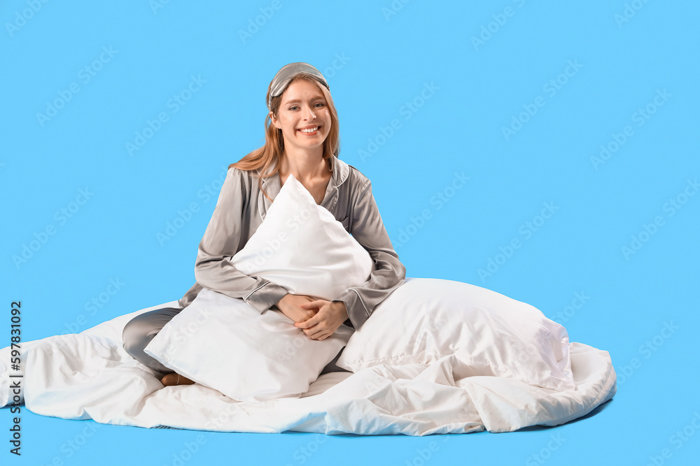 Young woman in pajamas with pillows and blanket on blue background