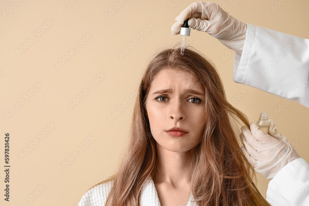 Cosmetologist applying serum for hair growth on young woman against beige background, closeup