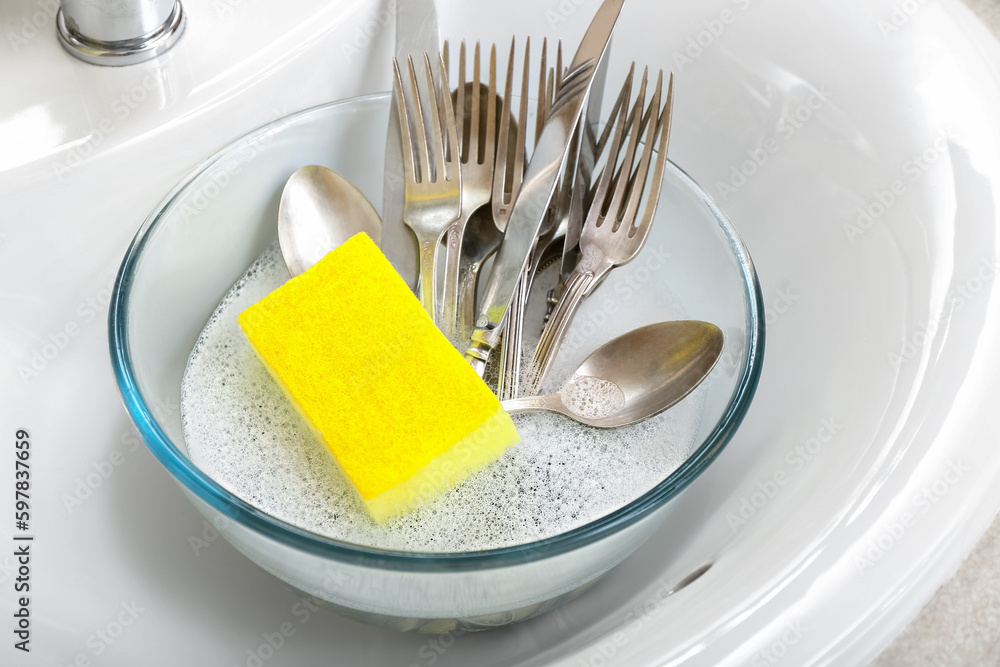 Bowl with cutlery and cleaning sponge in sink, closeup