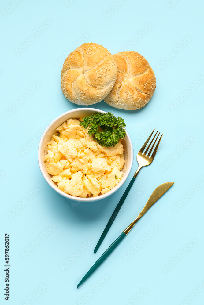 Bowl of scrambled eggs and buns with cutlery on blue background