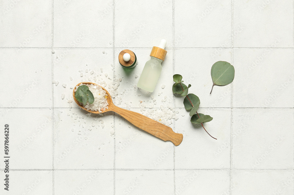 Cosmetic oil bottles with wooden spoon, eucalyptus branch and sea salt on tile background