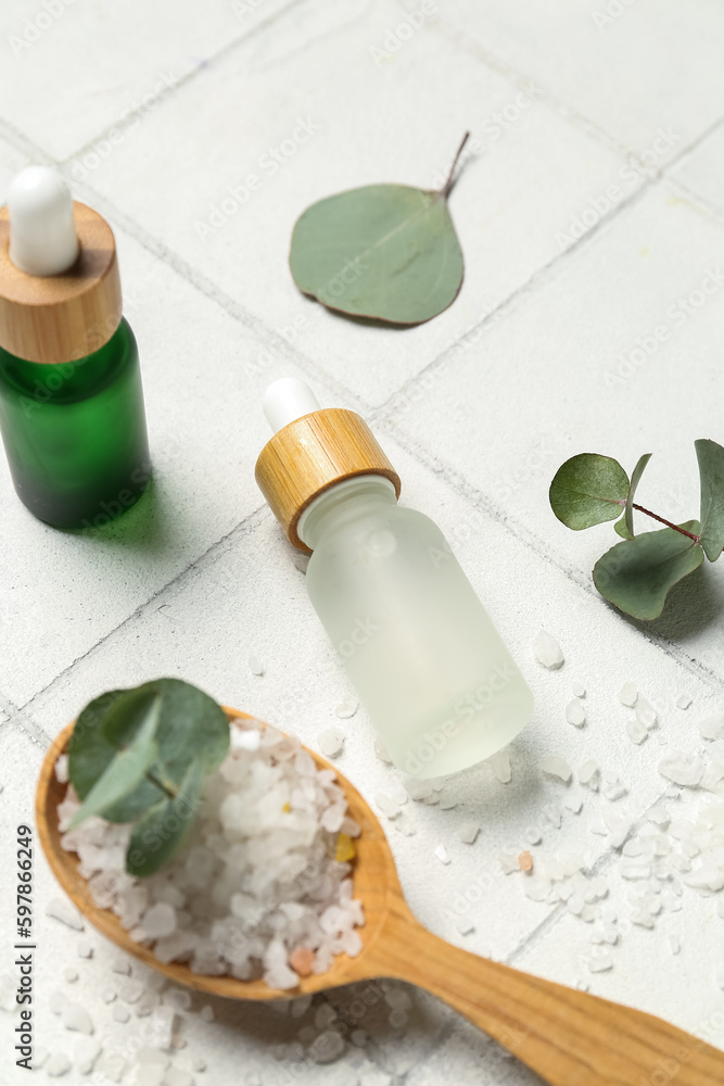 Cosmetic oil bottles with wooden spoon, eucalyptus branch and sea salt on tile background