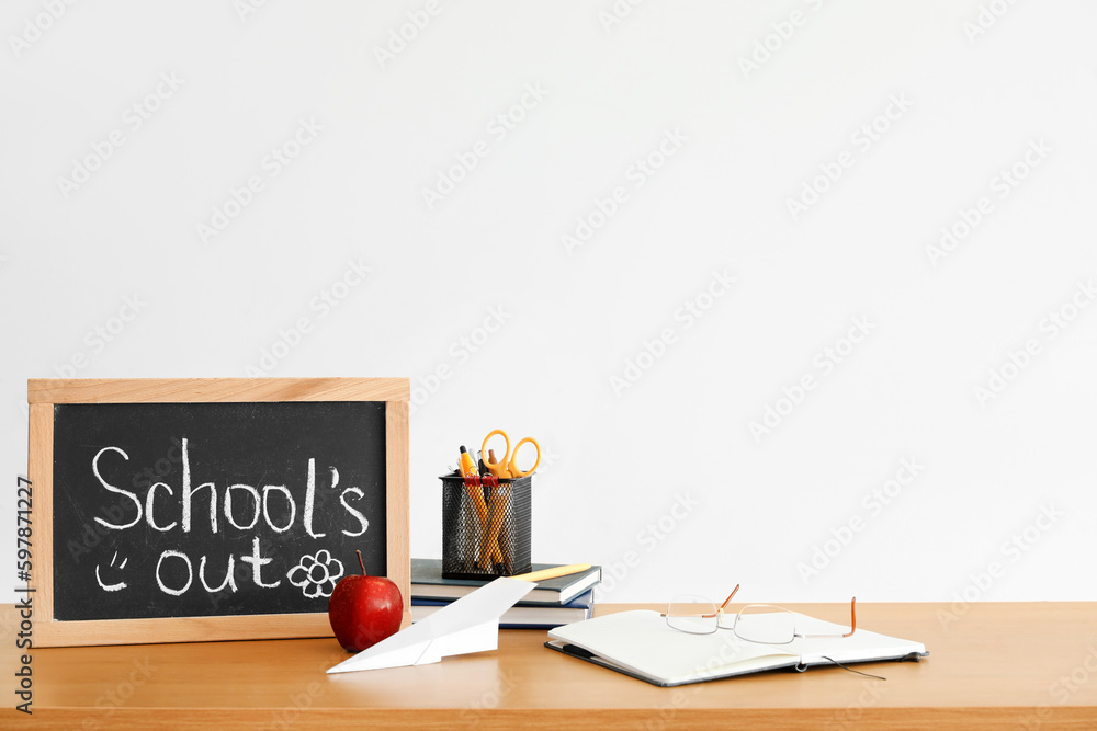 Chalkboard with text SCHOOLS OUT, apple, paper plane and stationery on table near light wall