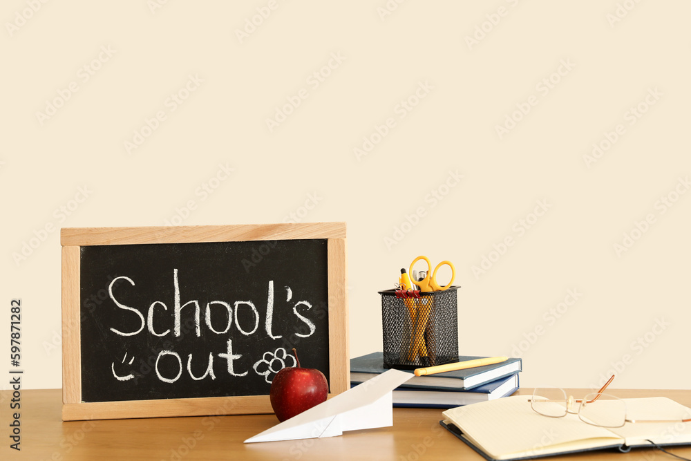 Chalkboard with text SCHOOLS OUT, apple, paper plane and stationery on table near light wall