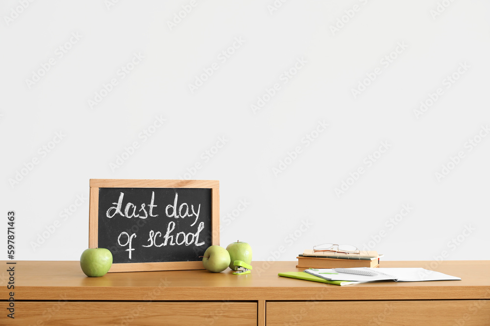 Chalkboard with text LAST DAY OF SCHOOL, apples and notebooks on table near light wall