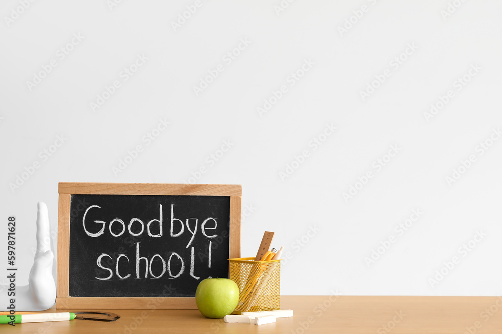 Chalkboard with text GOODBYE SCHOOL, apple and stationery on table near light wall