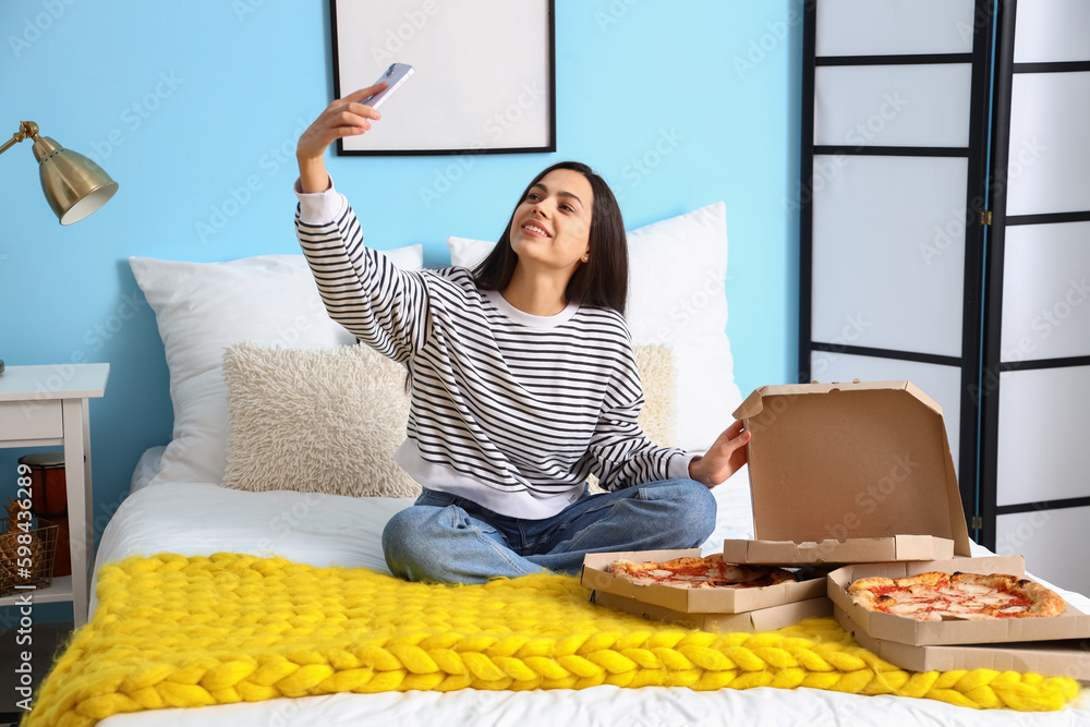 Young woman with boxes of tasty pizza taking selfie in bedroom