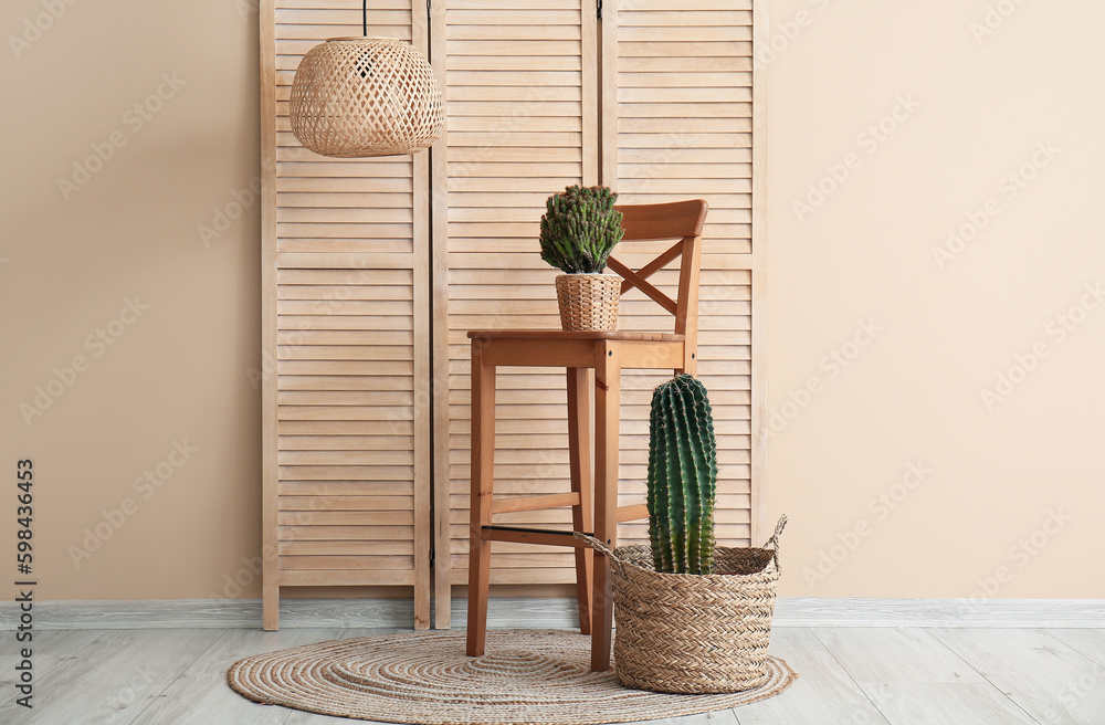 Different cacti in pots, chair and folding screen near beige wall