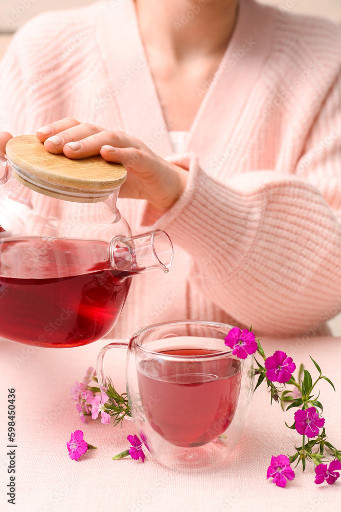 Woman pouring hot tea from teapot into cup with delicate flowers on pink table