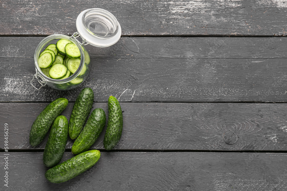 Jar with cut and whole cucumbers on dark wooden background