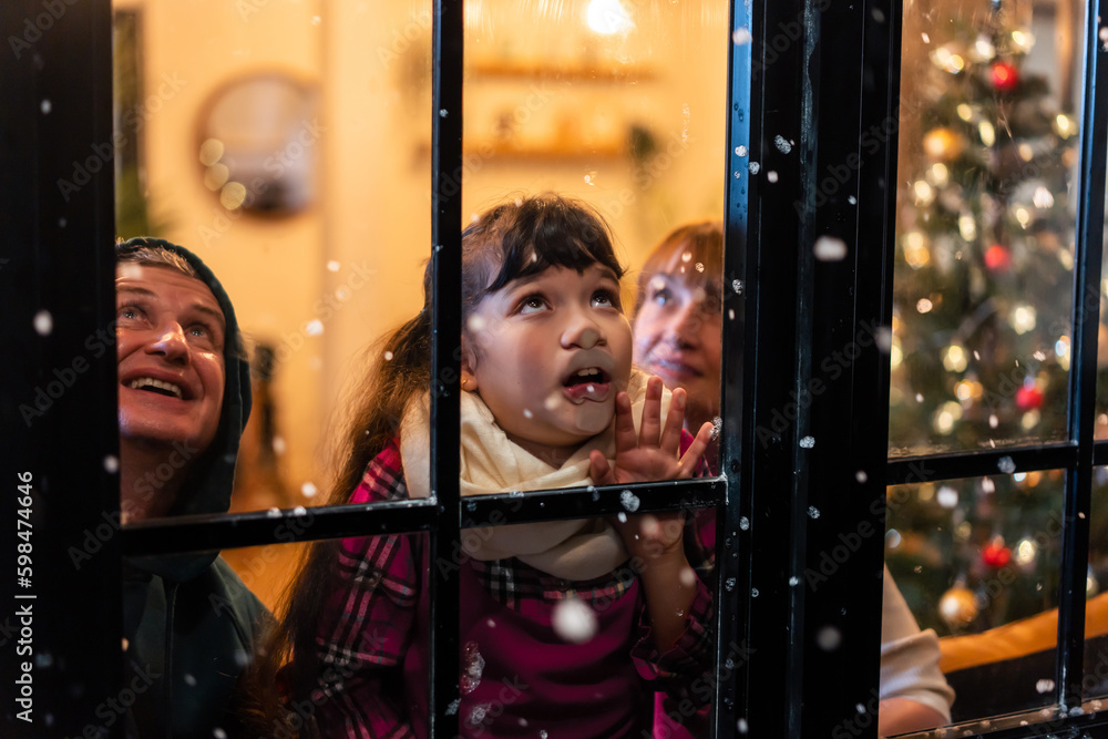 Adorable child looking at the window and first snow flakes with family. 