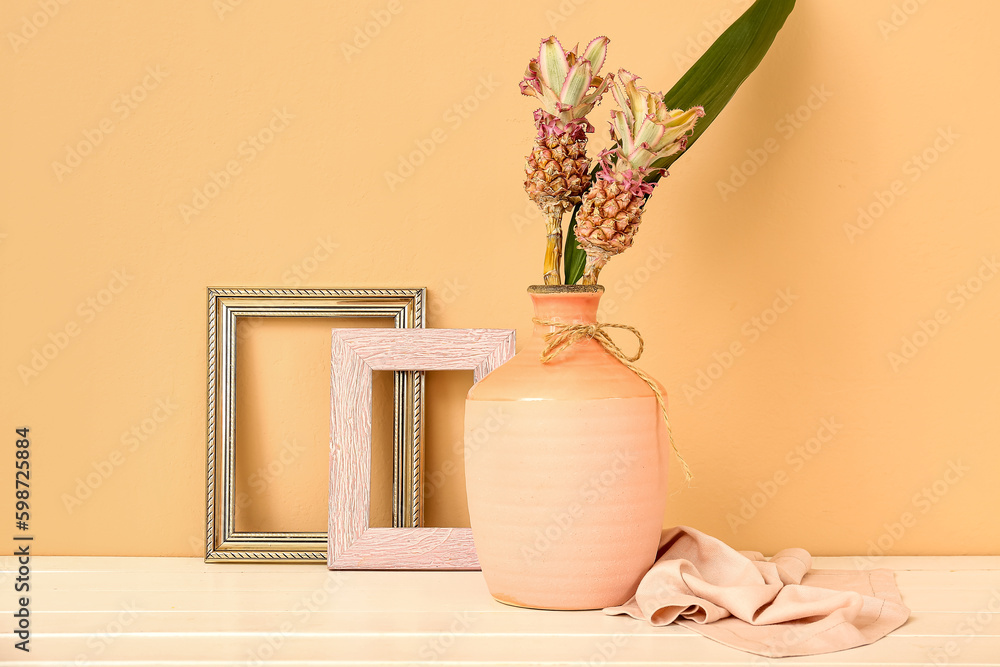 Vase with decorative pineapples and frames on table near beige wall