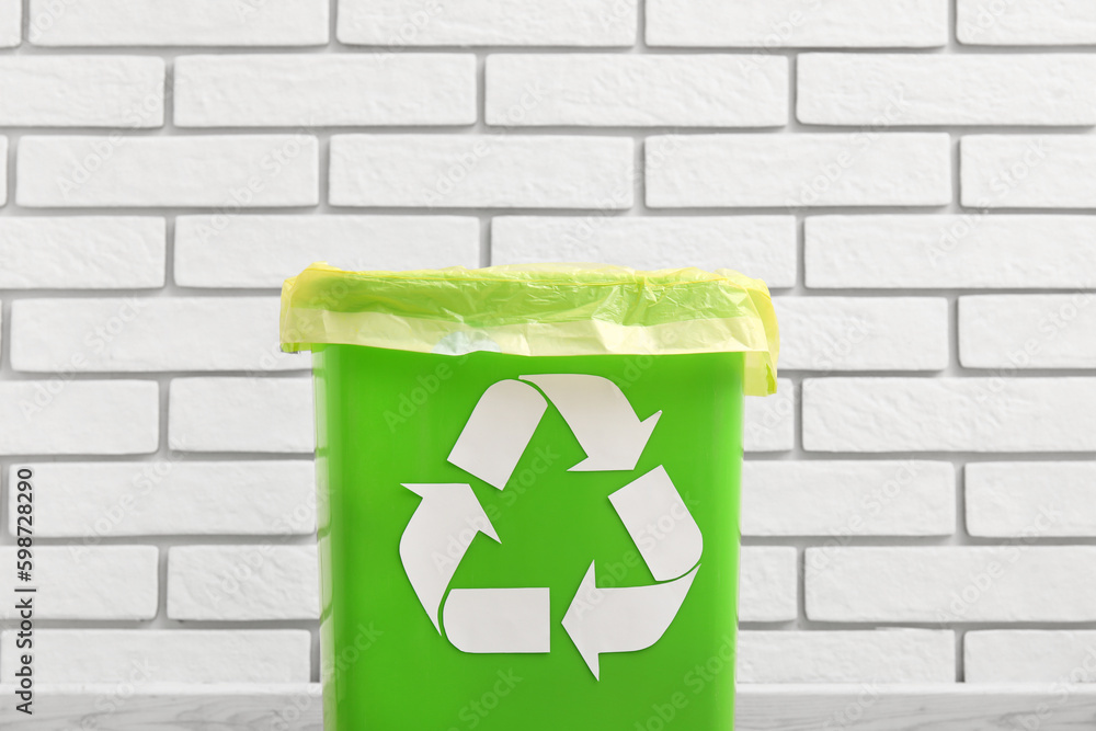 Trash bin with recycling symbol near white brick wall