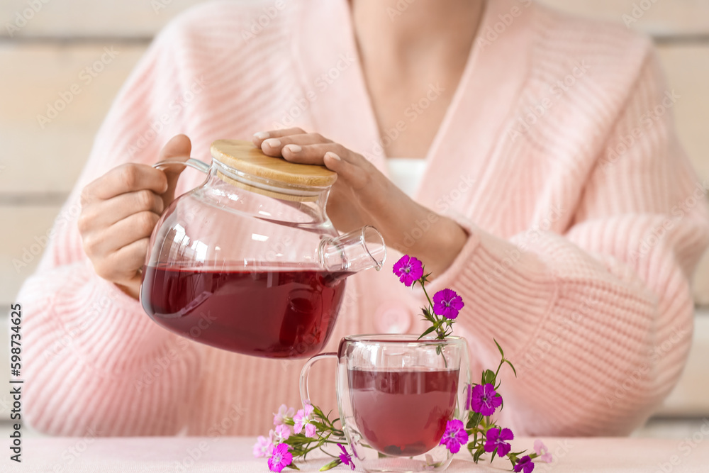 Woman pouring hot tea from teapot into cup with delicate flowers on pink table