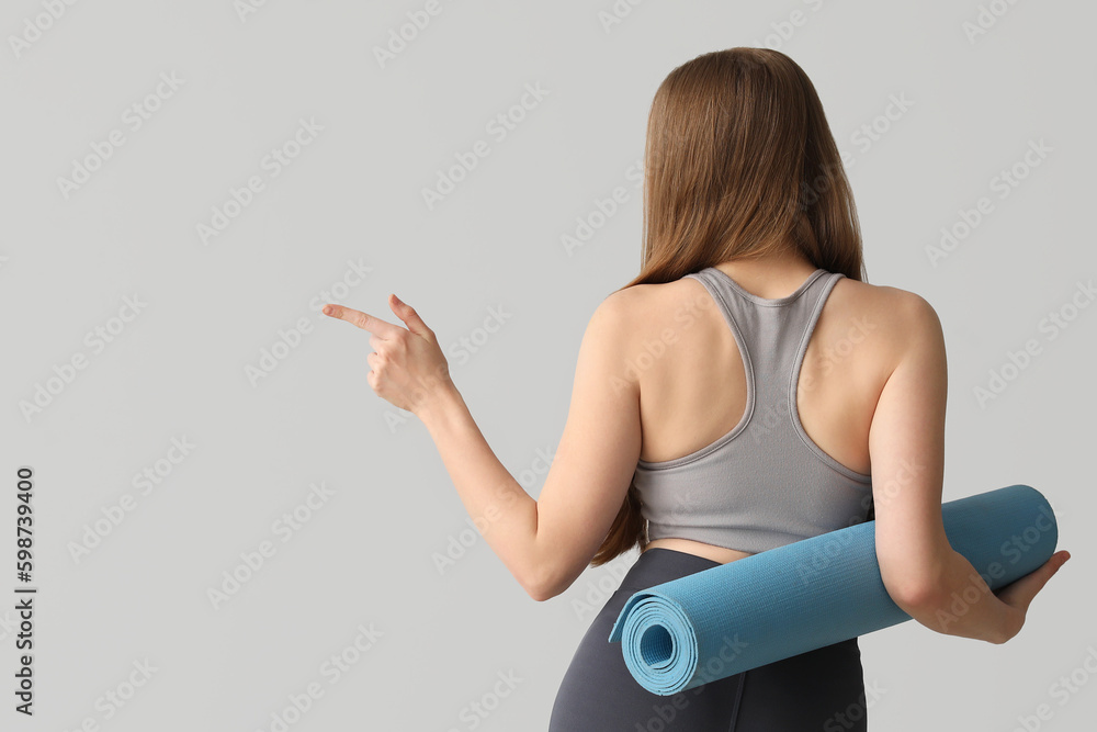 Sporty young woman with yoga mat pointing at something on grey background