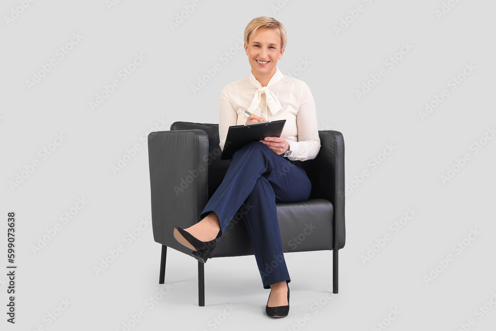 Mature businesswoman with clipboard sitting in armchair against grey background