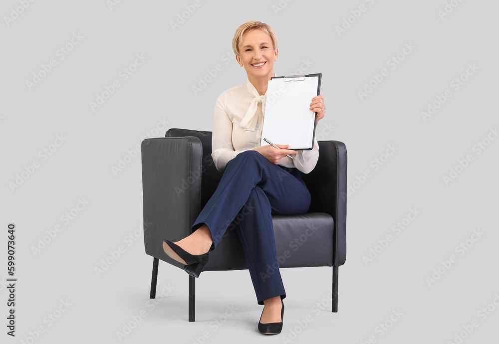 Mature businesswoman with clipboard sitting in armchair against grey background