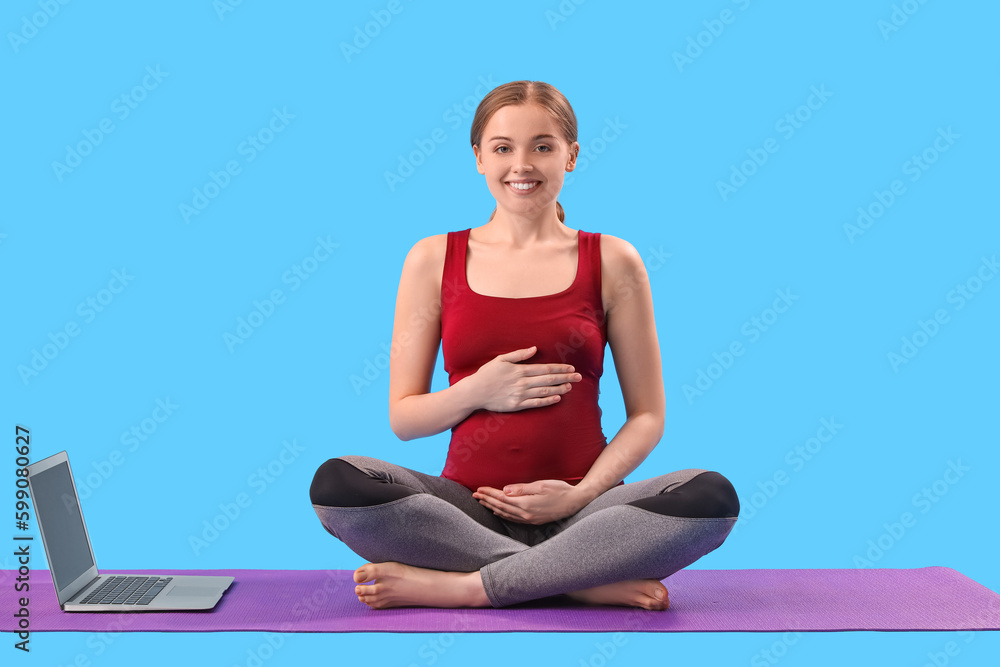 Pregnant young woman with laptop doing yoga on blue background