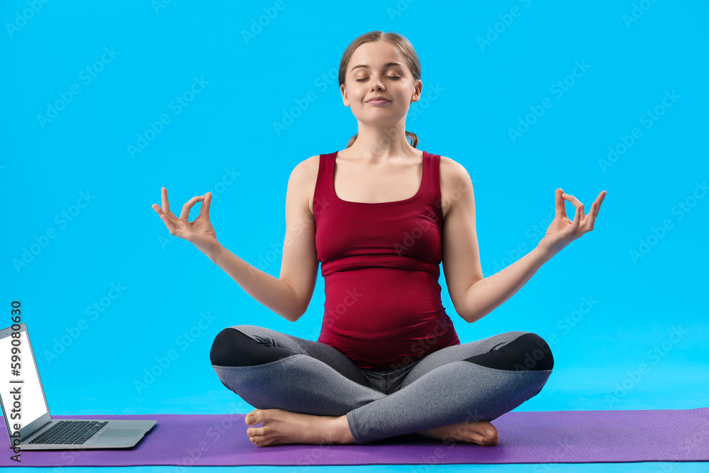 Pregnant young woman with laptop doing yoga on blue background