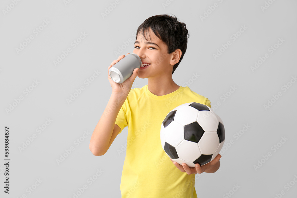 Little boy with soccer ball drinking soda on grey background