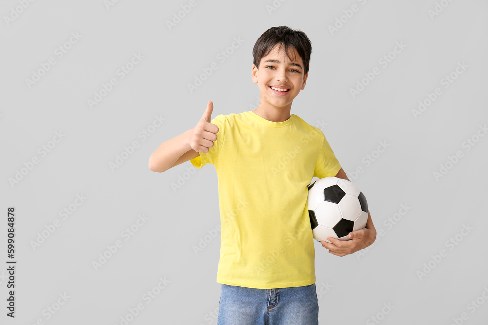 Little boy with soccer ball showing thumb-up on grey background