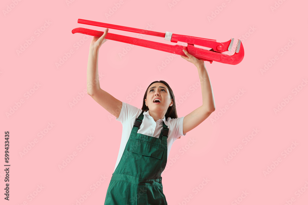 Worried female plumber with big pipe wrench on pink background