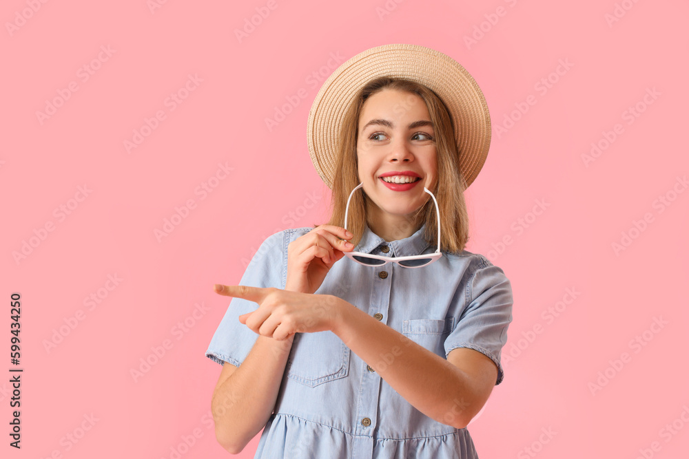 Young woman in hat with sunglasses pointing at something on pink background