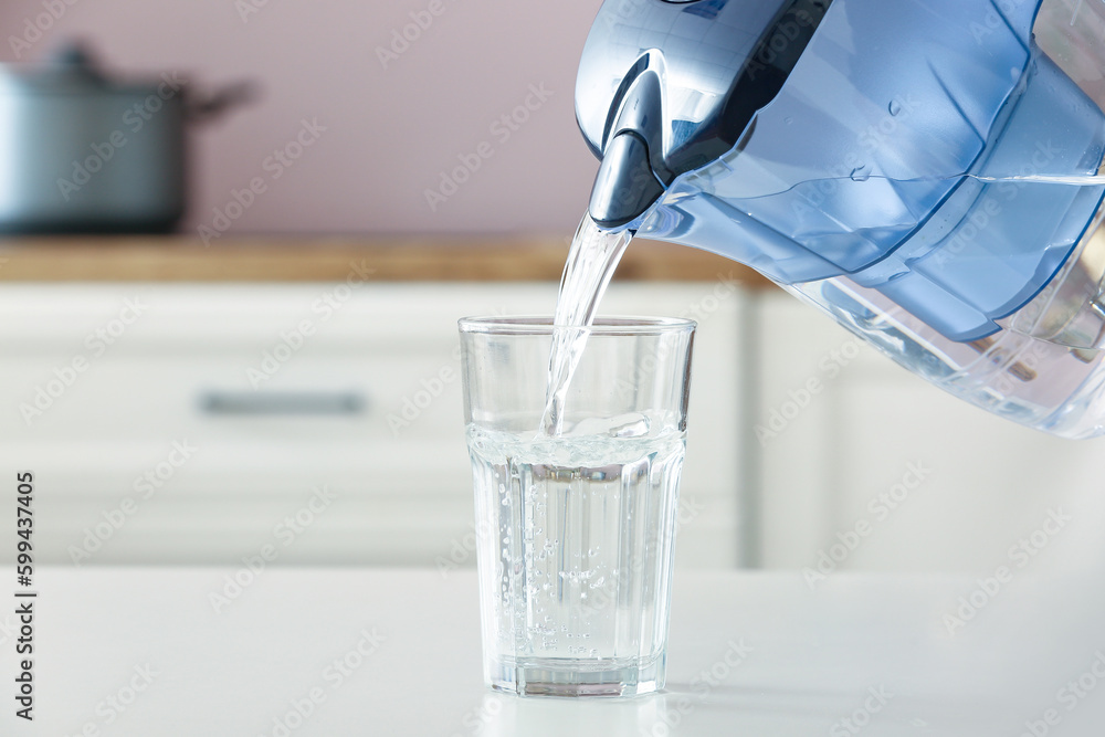 Pouring of water from filter jug into glass on table in kitchen