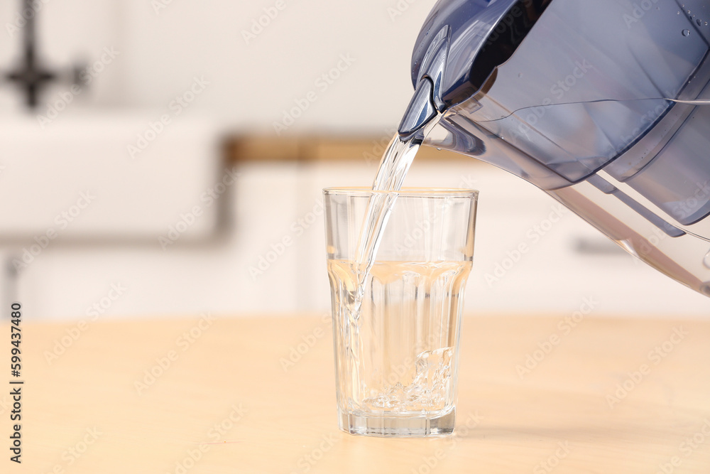 Pouring of pure water from filter jug into glass on table in kitchen