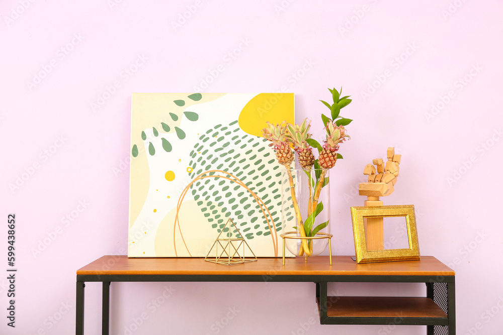 Vase with baby pineapples, painting and decor on table near lilac wall