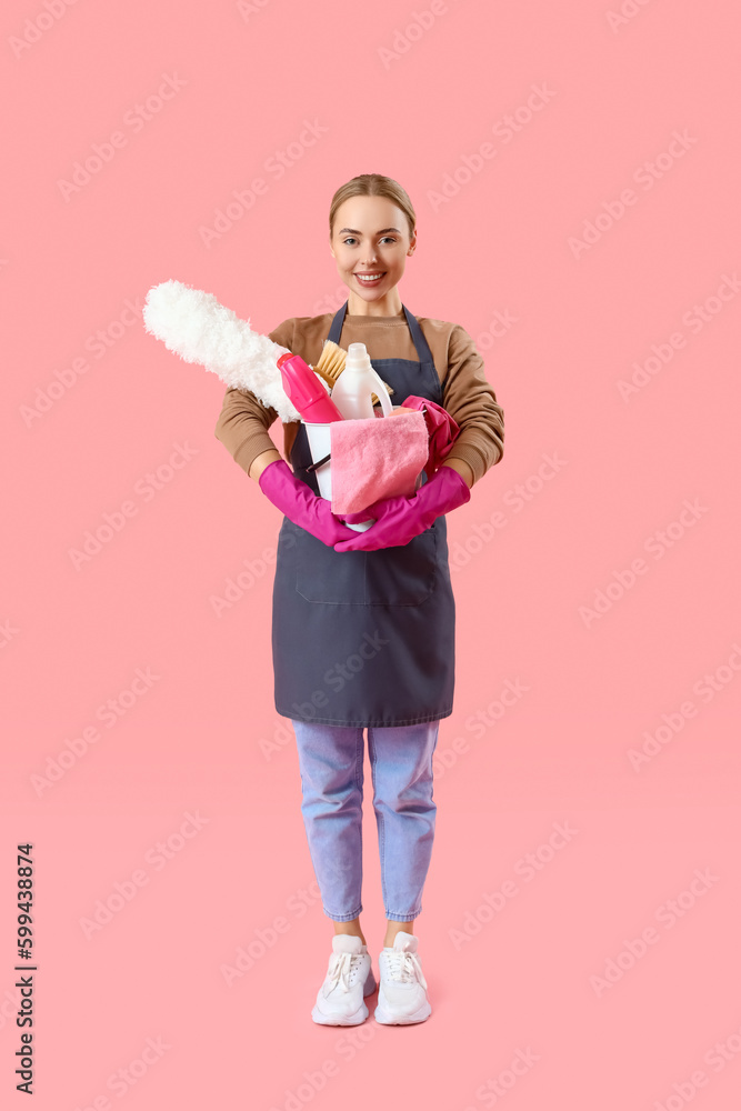 Young woman with bucket of cleaning supplies on pink background