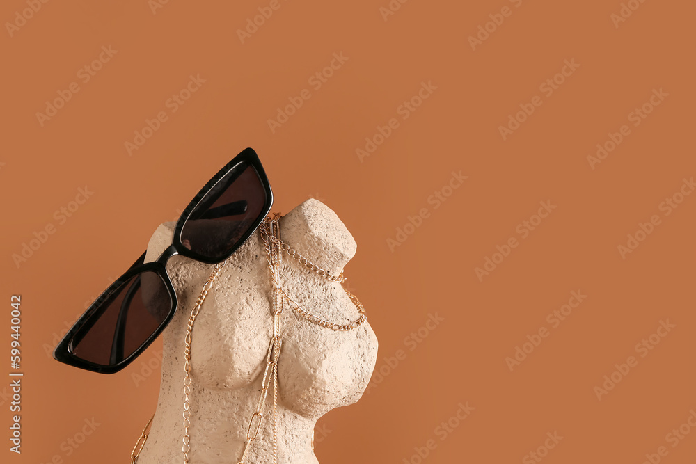 Statuette with stylish sunglasses and jewelry on brown background, closeup